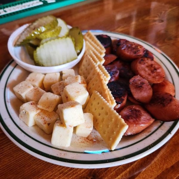 A plate of Ash's Irish Pub's cheese cubes, crackers, grilled sausage, and pickles.